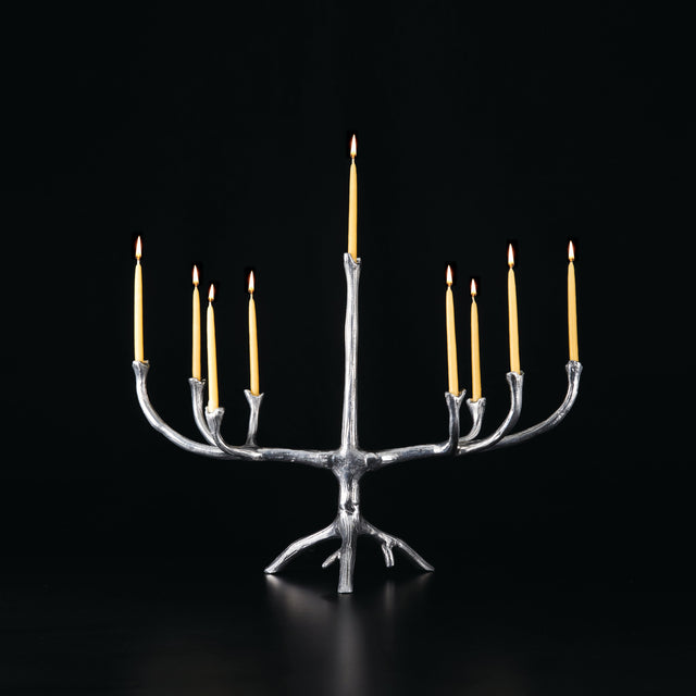 A rustic silver tree-branch style candle holder with nine arms, holding lit yellow taper candles, set against a solid black background.